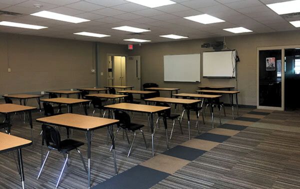 Activity Room A classroom-style activity room with rows of wooden desks and black chairs, whiteboards mounted on the wall, and overhead fluorescent lighting. The room features a striped carpet floor and an open door leading to another space.