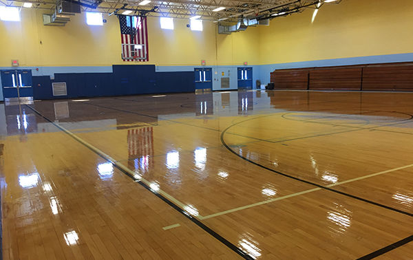 Gymnasium A spacious gymnasium with a polished wooden floor, marked with basketball court lines. The walls are painted yellow and blue, and an American flag hangs at the far end. The gym features bleacher seating on one side and several doors leading to other areas.